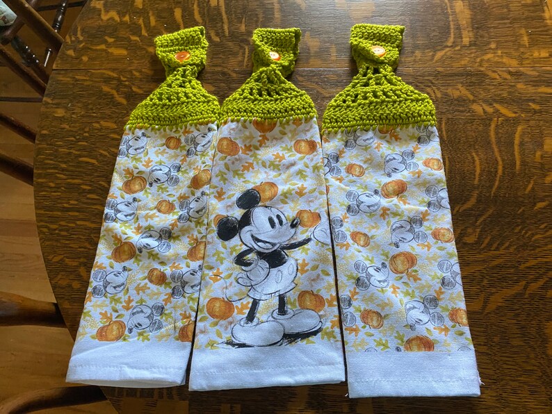 Op de afbeelding: Drie witte keukendoeken met een groene gehaakte bovenkant en een knoop. De handdoeken hebben een Mickey Mouse-figuur en een herfstthema met pompoenen en bladeren.