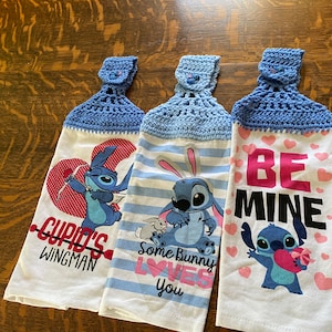 May include: Three blue and white kitchen towels with a crocheted blue hanging loop. The towels feature a cartoon character with different Valentine's Day themed messages. The first towel says "Cupid's Wingman", the second says "Some Bunny Loves You", and the third says "Be Mine".