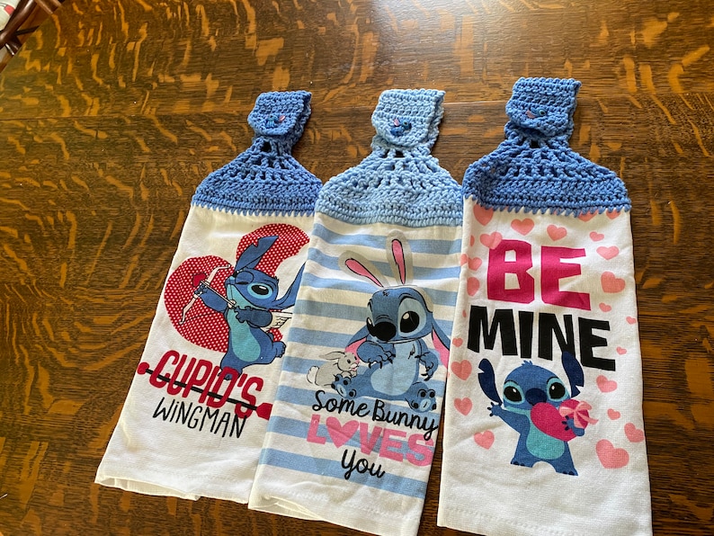 May include: Three blue and white kitchen towels with a crocheted blue hanging loop. The towels feature a cartoon character with different Valentine's Day themed messages. The first towel says "Cupid's Wingman", the second says "Some Bunny Loves You", and the third says "Be Mine".