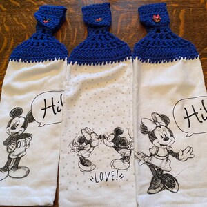 Op de afbeelding: Drie witte keukendoeken met blauwe gehaakte toppen en een Mickey Mouse en Minnie Mouse ontwerp. De handdoeken bevatten de tekst "Hi!" en "LOVE!" in zwart.
