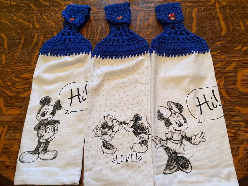 Op de afbeelding: Drie witte keukendoeken met blauwe gehaakte toppen en een Mickey Mouse en Minnie Mouse ontwerp. De handdoeken bevatten de tekst "Hi!" en "LOVE!" in zwart.