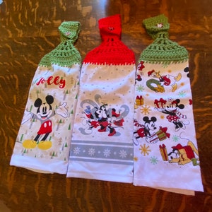 May include: Three white kitchen towels with Disney character designs. Each towel has a different design, including Mickey Mouse, Minnie Mouse, and other characters. The tops are crocheted in red and green, with a button closure. One towel says "Jolly".