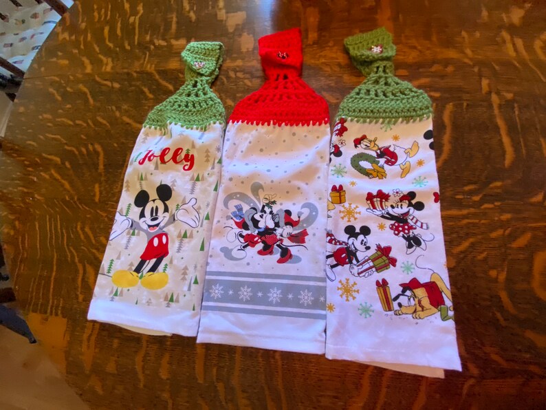 May include: Three white kitchen towels with Disney character designs. Each towel has a different design, including Mickey Mouse, Minnie Mouse, and other characters. The tops are crocheted in red and green, with a button closure. One towel says "Jolly".