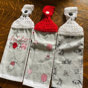 May include: Three hanging kitchen towels with a Winnie the Pooh theme. Two towels have a gray background with Pooh and balloon graphics. One towel says "hello POOH" with a red balloon. Each towel has a crocheted top.