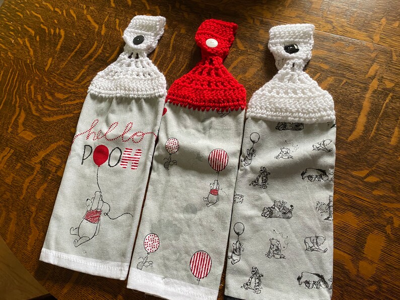 May include: Three hanging kitchen towels with a Winnie the Pooh theme. Two towels have a gray background with Pooh and balloon graphics. One towel says "hello POOH" with a red balloon. Each towel has a crocheted top.