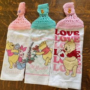 May include: Three Winnie the Pooh themed kitchen towels with crocheted hanging loops. The towels feature different designs: one with Winnie the Pooh and Piglet celebrating Easter, one with Winnie the Pooh smelling flowers, and one with Winnie the Pooh holding a love letter.
