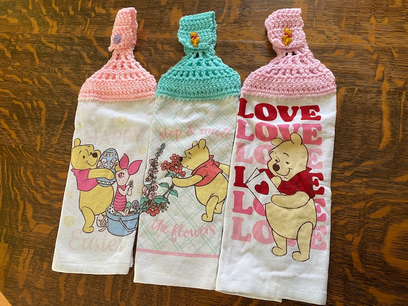 May include: Three Winnie the Pooh themed kitchen towels with crocheted hanging loops. The towels feature different designs: one with Winnie the Pooh and Piglet celebrating Easter, one with Winnie the Pooh smelling flowers, and one with Winnie the Pooh holding a love letter.