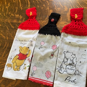 May include: Three Winnie the Pooh kitchen towels with crocheted tops. The first towel is white with red crocheted trim and features Winnie the Pooh with the text "Bee kind. Bee happy. Bee you". The second towel is grey with black crocheted trim and features Winnie the Pooh with red and white balloons. The third towel is white with red crocheted trim and features Winnie the Pooh with a pot of honey and the text "Hunny" and "Pooh".
