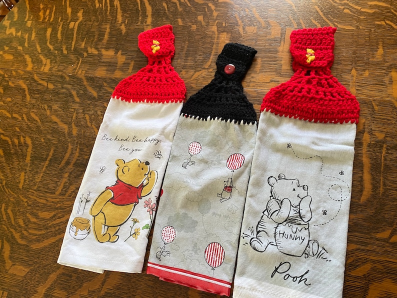 May include: Three Winnie the Pooh kitchen towels with crocheted tops. The first towel is white with red crocheted trim and features Winnie the Pooh with the text "Bee kind. Bee happy. Bee you". The second towel is grey with black crocheted trim and features Winnie the Pooh with red and white balloons. The third towel is white with red crocheted trim and features Winnie the Pooh with a pot of honey and the text "Hunny" and "Pooh".
