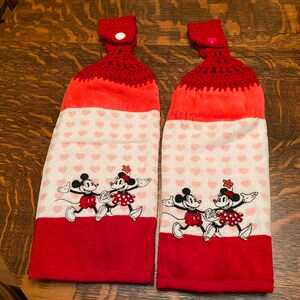 May include: Two red and white kitchen towels with a crocheted top and a pink button. The towels feature a heart pattern and a Mickey Mouse and Minnie Mouse design. The text on the towels reads "Happy Valentine's Day".