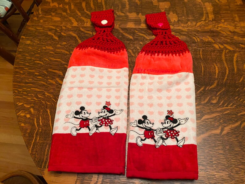 May include: Two red and white kitchen towels with a crocheted top and a pink button. The towels feature a heart pattern and a Mickey Mouse and Minnie Mouse design. The text on the towels reads "Happy Valentine's Day".