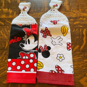 May include: Two red and white kitchen towels with a crocheted loop at the top. One towel features a large image of Minnie Mouse with the word "Minnie" printed on the towel. The other towel features a white background with a repeating pattern of Minnie Mouse's head, gloves, and polka dots.