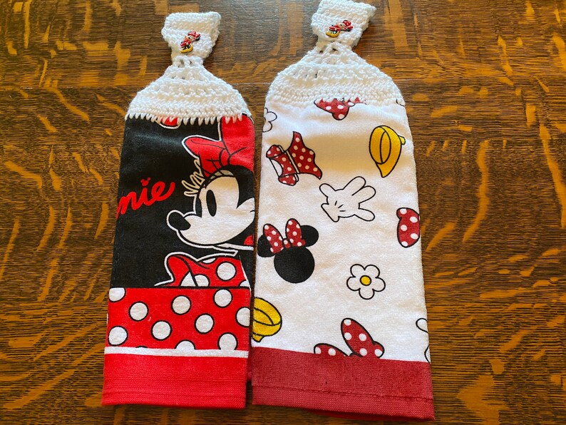 May include: Two red and white kitchen towels with a crocheted loop at the top. One towel features a large image of Minnie Mouse with the word "Minnie" printed on the towel. The other towel features a white background with a repeating pattern of Minnie Mouse's head, gloves, and polka dots.