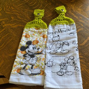 May include: Two white kitchen towels with green crocheted hanging loops. One towel features a black and white illustration of Mickey Mouse with pumpkins and fall leaves. The other towel has a black and white sketch of Mickey Mouse with the text "Mickey Mouse" and "Exaggerated Chest".