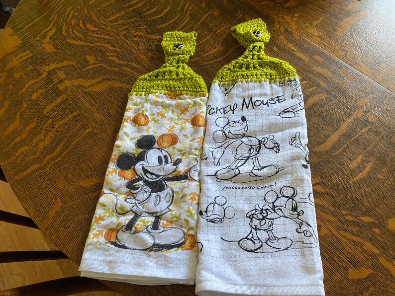 May include: Two white kitchen towels with green crocheted hanging loops. One towel features a black and white illustration of Mickey Mouse with pumpkins and fall leaves. The other towel has a black and white sketch of Mickey Mouse with the text "Mickey Mouse" and "Exaggerated Chest".