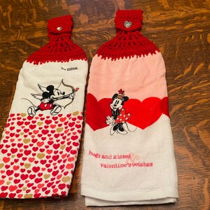 May include: Two red and white kitchen towels with a crocheted red hanging loop. One towel features Mickey Mouse as Cupid with the text "be mine." The other towel features Minnie Mouse with the text "hugs and kisses valentine's wishes."