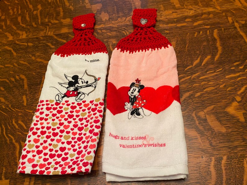 May include: Two red and white kitchen towels with a crocheted red hanging loop. One towel features Mickey Mouse as Cupid with the text "be mine." The other towel features Minnie Mouse with the text "hugs and kisses valentine's wishes."