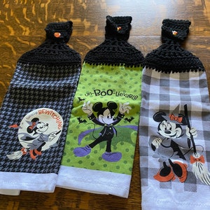 May include: Three Halloween-themed hanging kitchen towels. Each towel features a different design with a black crocheted top and a decorative button. The towels have images of Mickey and Minnie Mouse in Halloween costumes, with the text "Totally Bewitching" and "un-Boo-lievable".