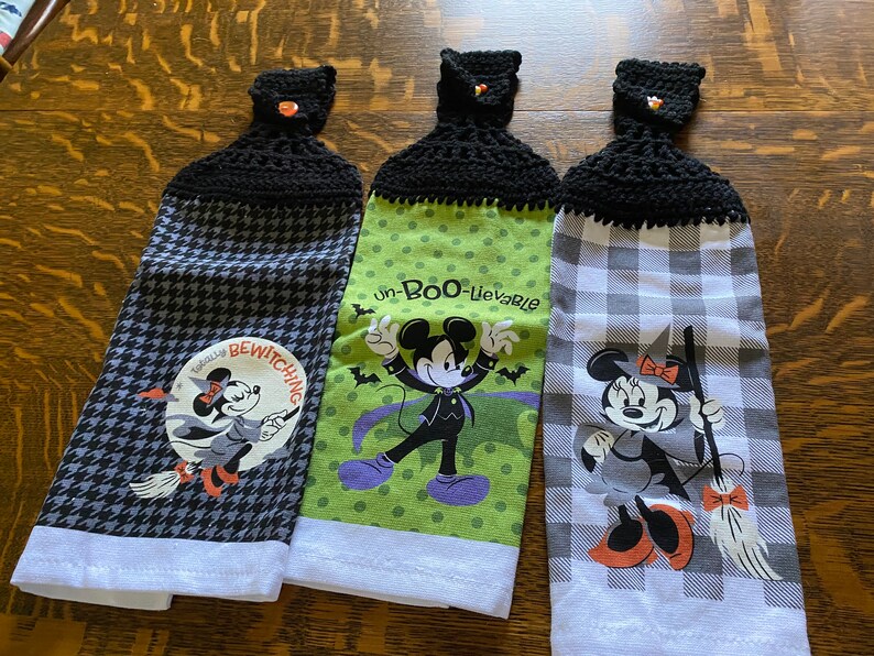 May include: Three Halloween-themed hanging kitchen towels. Each towel features a different design with a black crocheted top and a decorative button. The towels have images of Mickey and Minnie Mouse in Halloween costumes, with the text "Totally Bewitching" and "un-Boo-lievable".