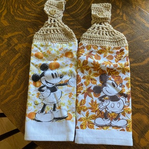 May include: Two white kitchen towels with a fall theme. Each towel features a cartoon image of Mickey Mouse and a pattern of orange, yellow, and brown leaves. The towels have crocheted loops at the top.