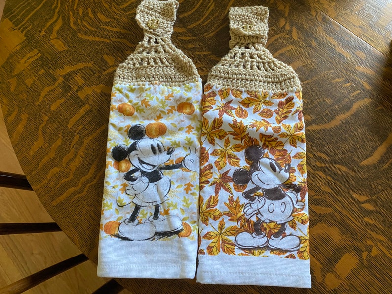 May include: Two white kitchen towels with a fall theme. Each towel features a cartoon image of Mickey Mouse and a pattern of orange, yellow, and brown leaves. The towels have crocheted loops at the top.