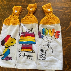 Op de afbeelding: Set van drie keukendoeken met een gele gehaakte bovenkant. Elke handdoek heeft een ander ontwerp met een cartoon Snoopy en regenboogkleuren. De ontwerpen omvatten Snoopy die een regenboogballon vasthoudt met de tekst "LOVE", Snoopy die op een regenboog hondenhok zit met de tekst "be happy", en Snoopy met een regenboogkwast.