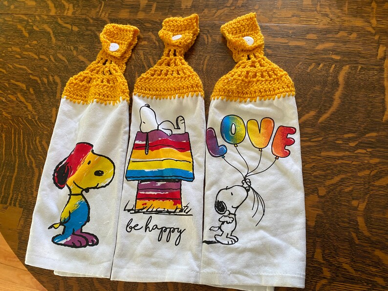 Op de afbeelding: Set van drie keukendoeken met een gele gehaakte bovenkant. Elke handdoek heeft een ander ontwerp met een cartoon Snoopy en regenboogkleuren. De ontwerpen omvatten Snoopy die een regenboogballon vasthoudt met de tekst "LOVE", Snoopy die op een regenboog hondenhok zit met de tekst "be happy", en Snoopy met een regenboogkwast.