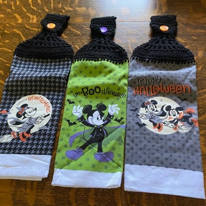 May include: Three Halloween-themed hanging kitchen towels. Each towel has a black crocheted top with a button. The towels feature Mickey and Minnie Mouse in Halloween costumes, with phrases like "Bewitching" and "Happy Halloween".