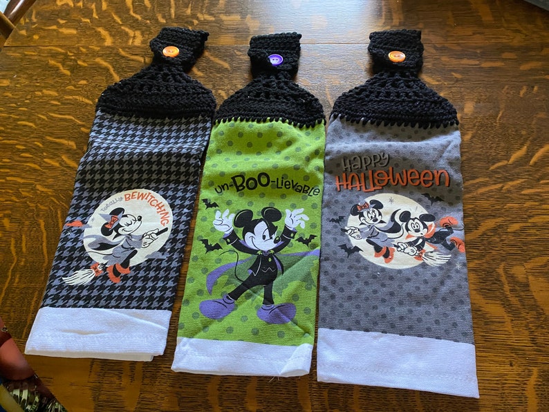May include: Three Halloween-themed hanging kitchen towels. Each towel has a black crocheted top with a button. The towels feature Mickey and Minnie Mouse in Halloween costumes, with phrases like "Bewitching" and "Happy Halloween".
