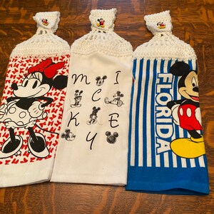 Op de afbeelding: Drie witte keukendoeken met gecrochete lussen. De eerste handdoek heeft Minnie Mouse in een rode jurk met witte stippen op een rode hartjesachtergrond. De tweede handdoek heeft Mickey Mouse-letters en -afbeeldingen. De derde handdoek heeft een blauw-wit gestreepte achtergrond met Mickey Mouse en het woord "FLORIDA".