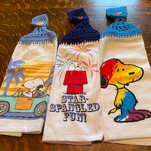 Op de afbeelding: Drie witte keukendoeken met gebreide blauwe en witte ophanglussen. Elke handdoek heeft een ander Peanuts-personageontwerp. De eerste handdoek toont Snoopy die een blauwe auto bestuurt met Woodstock naast hem. De tweede handdoek toont Snoopy die op zijn hondenhok zit met vuurwerk op de achtergrond. De tekst "Star-Spangled Fun!" is onder de afbeelding gedrukt. De derde handdoek toont Snoopy met een regenboogkleurige kop en lichaam.