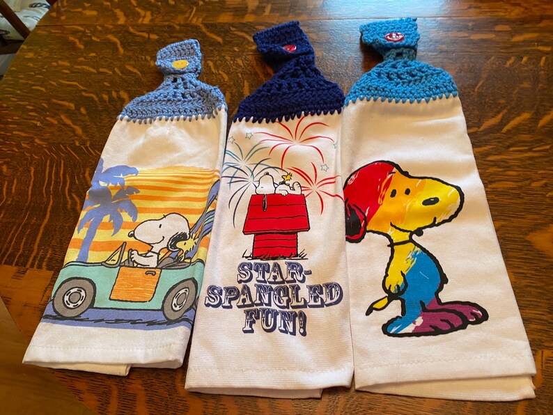 Op de afbeelding: Drie witte keukendoeken met gebreide blauwe en witte ophanglussen. Elke handdoek heeft een ander Peanuts-personageontwerp. De eerste handdoek toont Snoopy die een blauwe auto bestuurt met Woodstock naast hem. De tweede handdoek toont Snoopy die op zijn hondenhok zit met vuurwerk op de achtergrond. De tekst "Star-Spangled Fun!" is onder de afbeelding gedrukt. De derde handdoek toont Snoopy met een regenboogkleurige kop en lichaam.