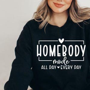 May include: Black sweatshirt with white text that reads "Homebody mode all day every day" with a heart symbol between "all day" and "every day".