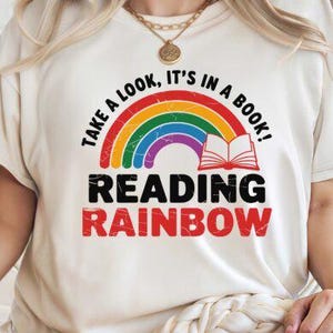 May include: Cream-colored t-shirt featuring a colorful rainbow design with the text "Take a look, it's in a book!" above "Reading Rainbow" in bold letters. The design includes an open book graphic. The shirt is a casual wear item.
