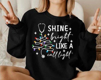 May include: Black sweatshirt with the text "Shine bright like a call light" in white. The design includes a stethoscope, Christmas lights, and stars. A festive and medical-themed design.