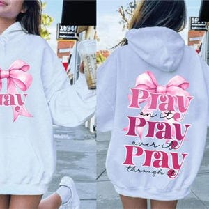 May include: White hoodie with a pink bow and the text "Pray" on the front. The back of the hoodie has a pink bow and the text "Play on it Play over it Play through it".