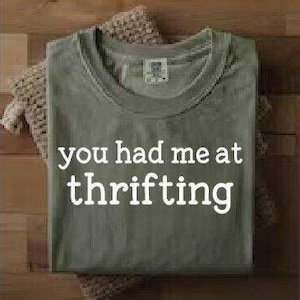May include: An olive green t-shirt, folded and displaying the white text "you had me at thrifting". It rests on a beige crocheted cloth, against a wooden backdrop. The shirt features a crew neck and short sleeves.
