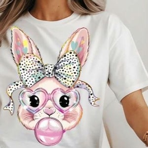 May include: White t-shirt with a colorful cartoon bunny wearing heart-shaped glasses and a polka dot bow. The bunny is blowing a pink bubble.