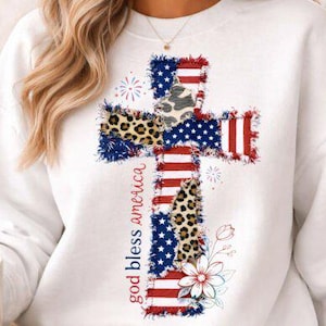 May include: White sweatshirt featuring a cross design with American flag elements, leopard print accents, and the text "god bless america" in red. The design includes stars, stripes, and floral details, creating a patriotic theme.