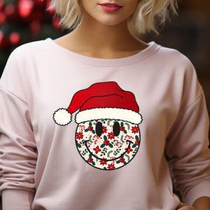 May include: A light pink long-sleeved sweatshirt featuring a smiley face design wearing a Santa hat. The smiley face is decorated with a floral pattern in red, white, and green. The background is blurred, with a Christmas tree visible.