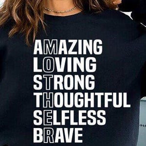 May include: Black sweatshirt with white text that reads "AMAZING LOVING STRONG THOUGHTFUL SELFLESS BRAVE"