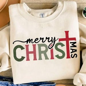 May include: Cream-colored sweatshirt with the words "merry CHRISTmas" in a festive design. The word "merry" is in black script, "CHRIS" is in green and red block letters, and "MAS" is in red with a cross.