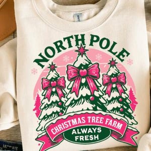May include: Cream-colored sweatshirt with a festive Christmas tree design. The graphic features three snow-covered trees with pink bows, the words "North Pole" and "Christmas Tree Farm Always Fresh" in green and pink.