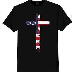 Puede incluir: Camiseta negra con un diseño de cruz de la bandera estadounidense desgastada. La cruz está en rojo, blanco y azul, con estrellas y rayas. La camiseta es de estilo cuello redondo clásico.