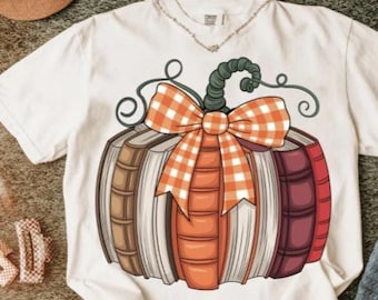 May include: White t-shirt featuring a pumpkin design made of books. The pumpkin has a green stem, an orange and white checkered bow, and is composed of books in shades of brown, orange, and red. A fall-themed graphic tee.