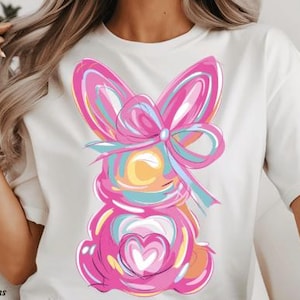 May include: White t-shirt with a colorful abstract bunny design. The bunny is pink, blue, yellow, and white with a large pink bow.