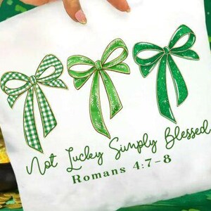 May include: White tote bag featuring three green bows, one with a gingham pattern. The text "Not Lucky Simply Blessed" and "Romans 4:7-8" are written in green script. The bag is held by a person.