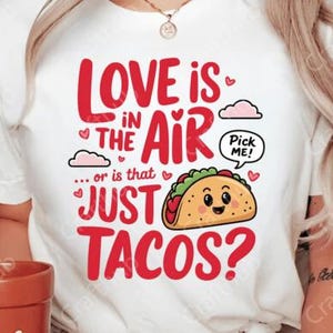 以下が含まれることがあります： 赤い文字で「Love is in the air... or is that just tacos?」と書かれた白いTシャツ。顔のある漫画のタコスと、その上に「Pick me!」と書かれた吹き出しがあります。