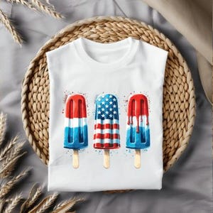 Può includere: T-shirt bianco con tre disegni di ghiaccioli ad acquerello rossi, bianchi e blu, a tema patriottico. I ghiaccioli sono su bastoncini di legno. La maglietta è esposta su un cesto intrecciato.