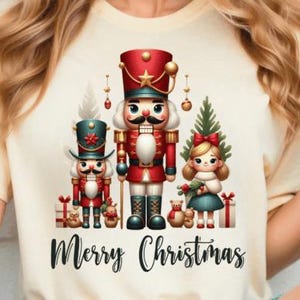 Könnte beinhalten: Cremefarbenes T-Shirt mit festlichem Weihnachtsdesign. Die Grafik zeigt einen Nussknacker, ein Mädchen und einen Weihnachtsbaum mit dem Schriftzug "Merry Christmas" in Schreibschrift. Geschenke und Teddybären sind ebenfalls enthalten.